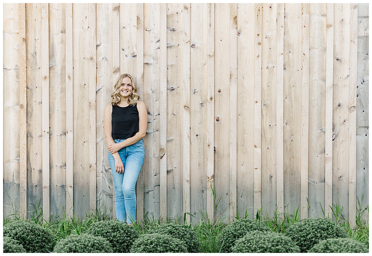 Girl smiling and leaning against a slatted wood wall with green bushes in front of her