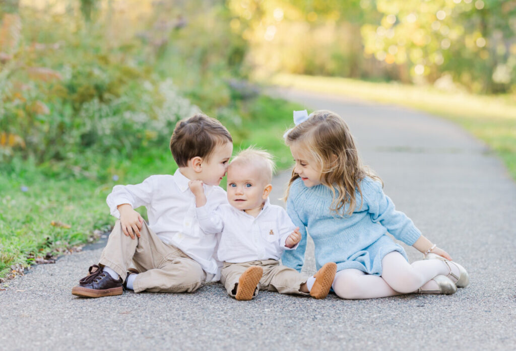 three children sitting on a path smiling at each other.